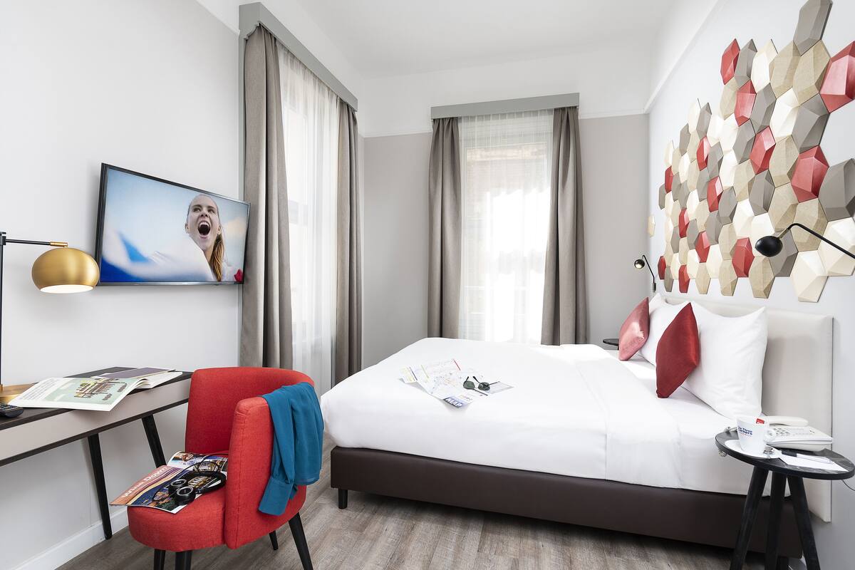 A modern hotel room with a bed, a desk, a chair, and a wall art piece.