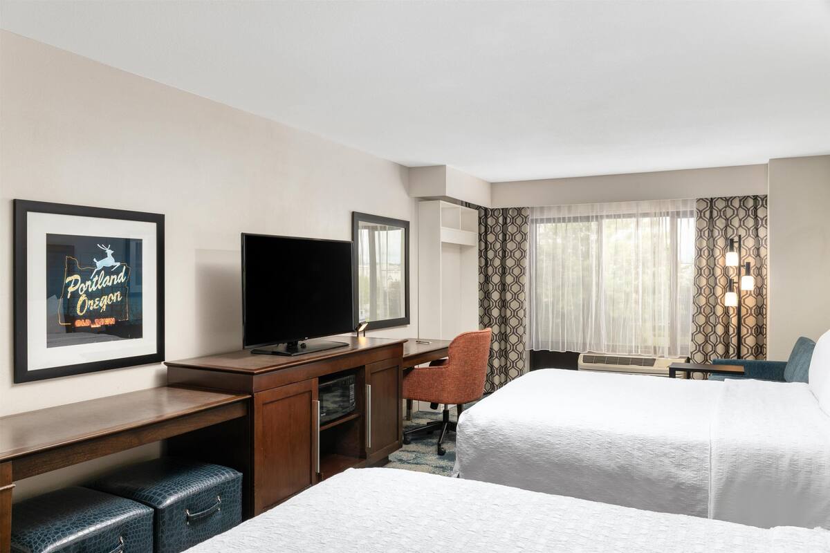 A hotel room with two beds, a desk with a flat-screen TV, a chair, a desk lamp, and a framed picture on the wall.
