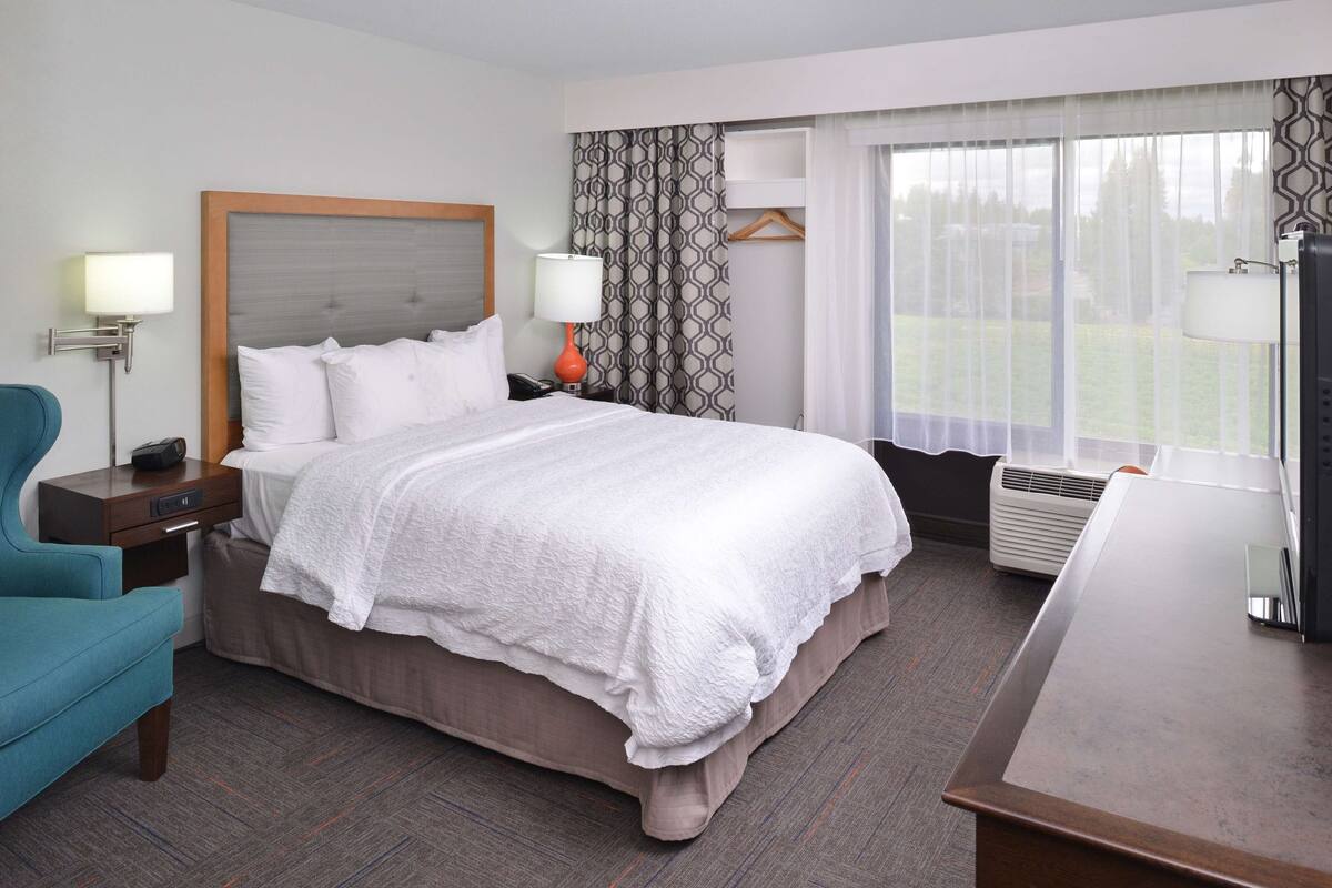 A hotel room with a bed, bedside table, lamp, chair, and a view of the outdoors.