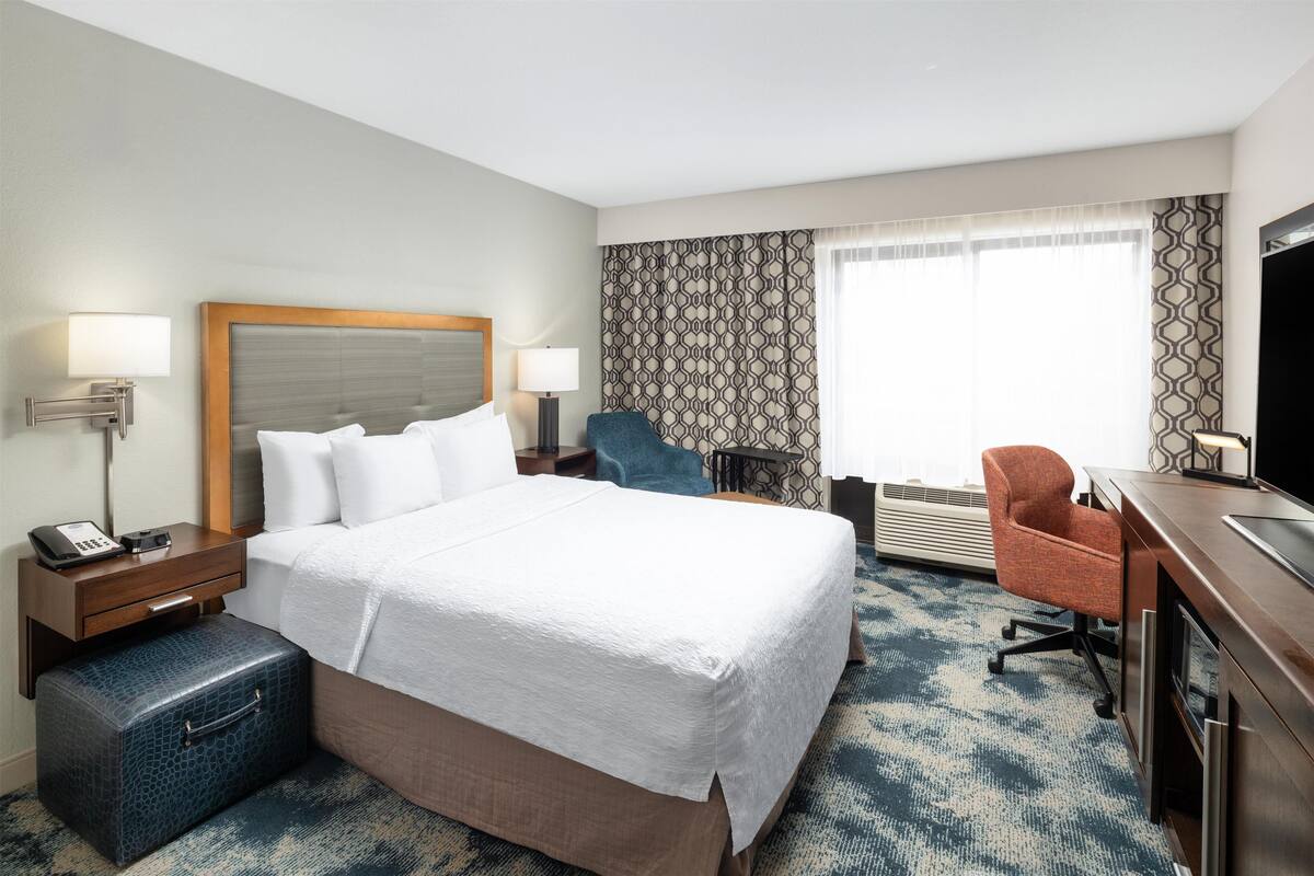 A hotel room with a large bed, a desk, a chair, a suitcase, and a television.