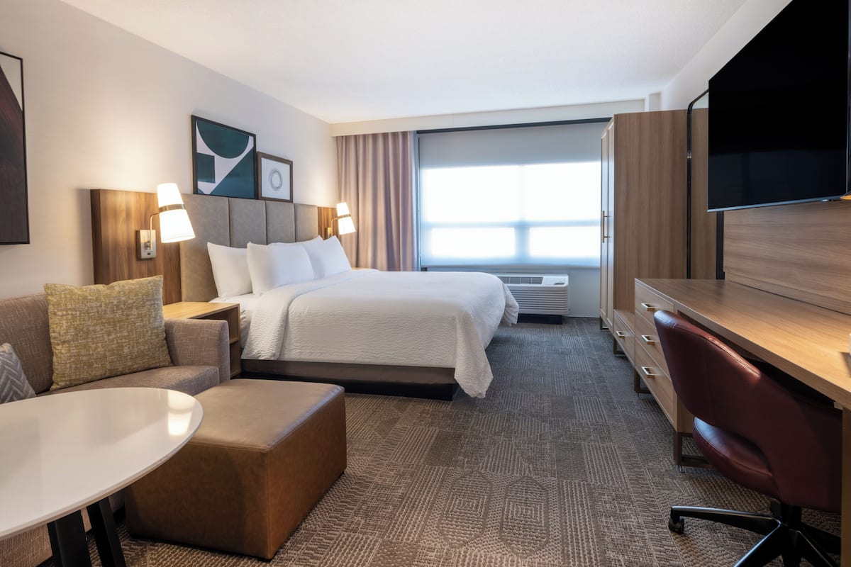 Une chambre d’hôtel comprenant un lit, un bureau, une chaise, une télévision, une fenêtre avec des rideaux et une lampe fixée au mur.