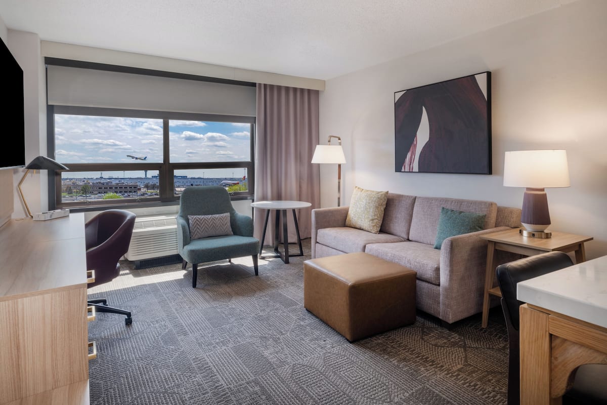 Une chambre d’hôtel avec une grande fenêtre, un canapé, un fauteuil, une petite table et une lampe.