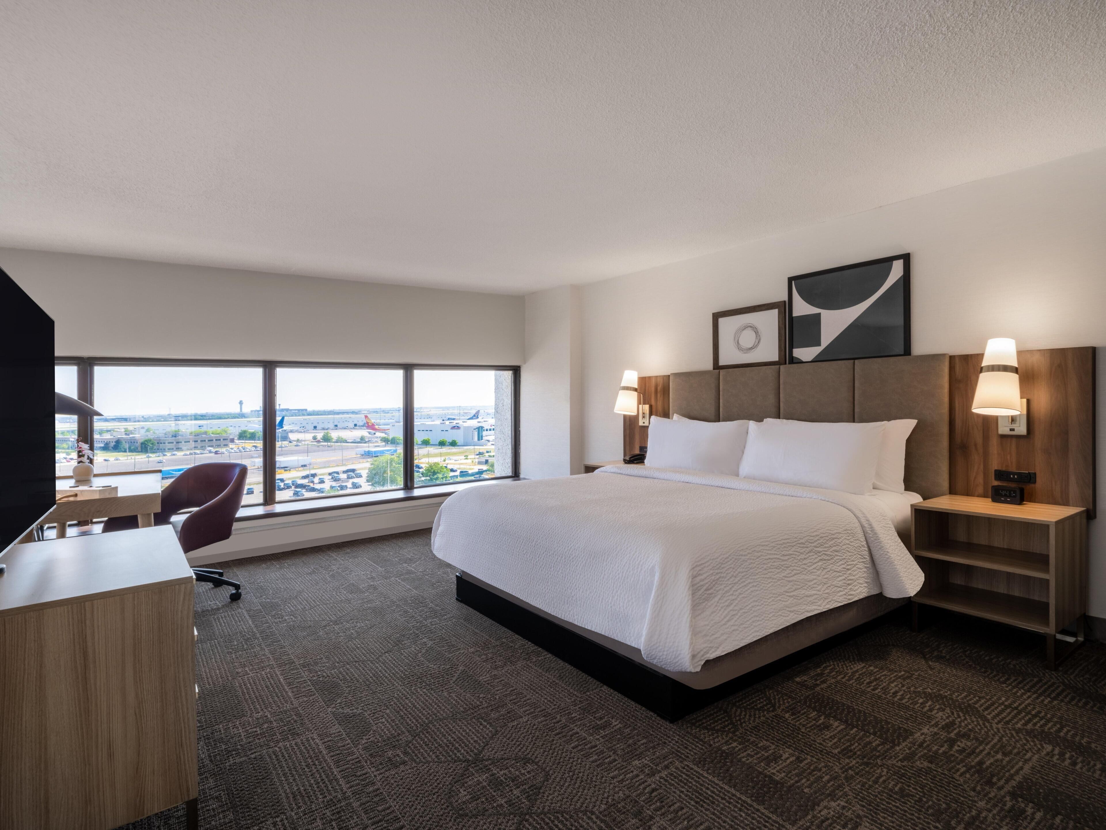 Une chambre d’hôtel avec un grand lit, un bureau et une chaise. Il y a une fenêtre donnant sur un aéroport et des immeubles.