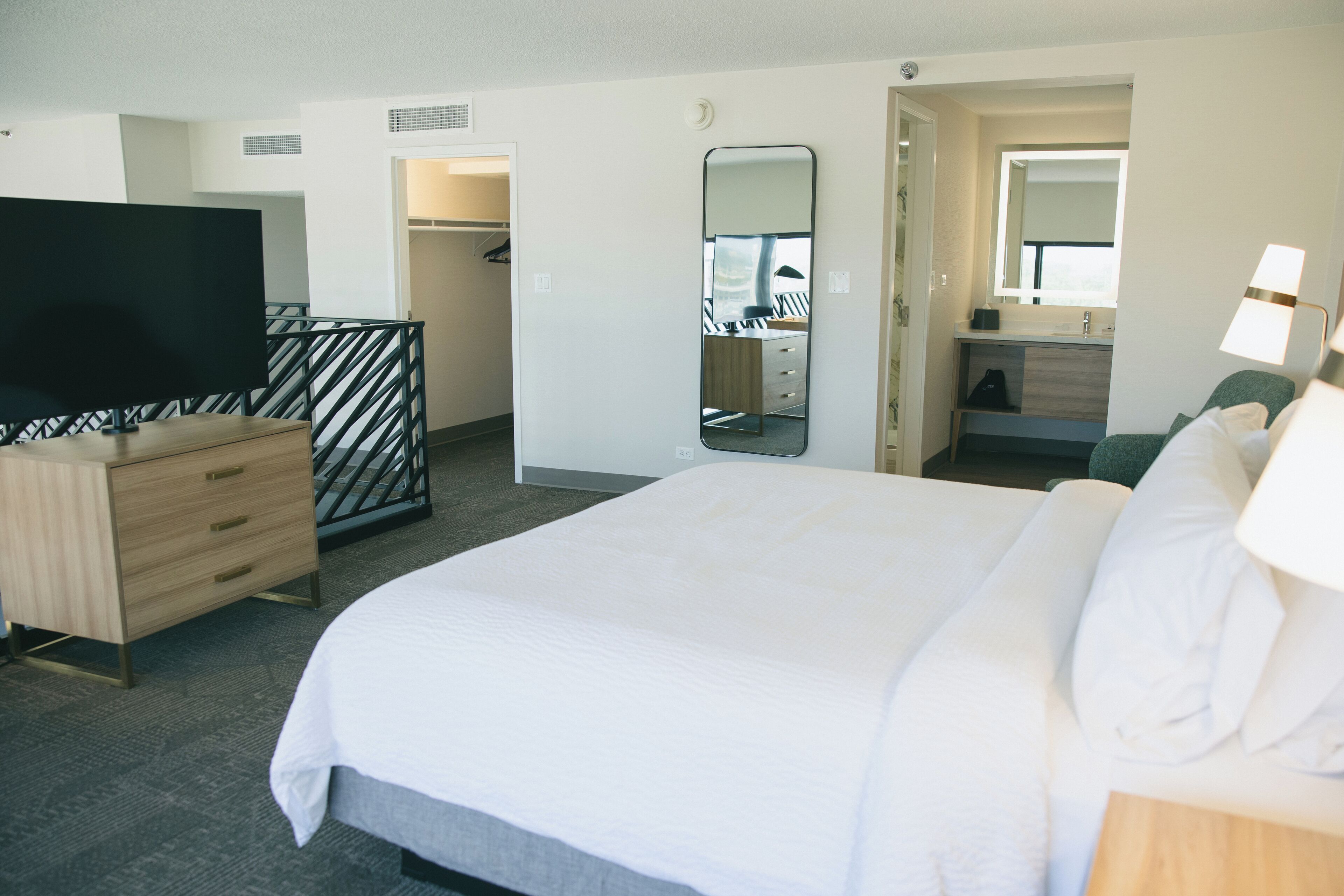 A hotel room with a large bed, a TV on a wooden dresser, a mirror, a desk with a chair, and a bathroom with a sink and mirror.