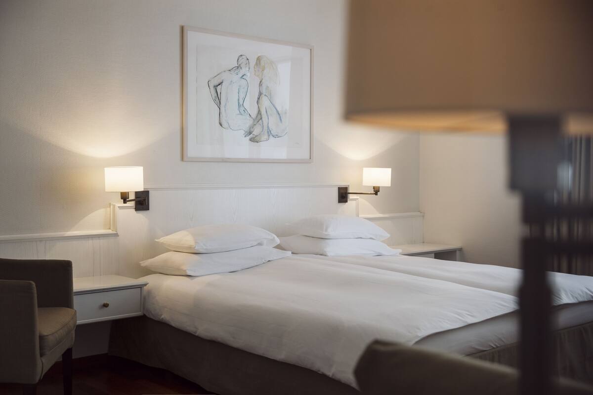 Habitación de hotel con cama doble, dos mesitas de noche, una silla y un cuadro enmarcado en la pared.