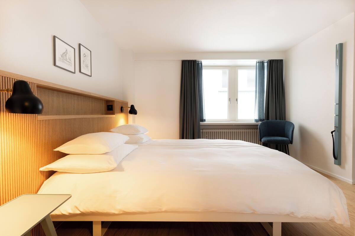 A hotel room with a large bed, a bedside table, a chair, and a window with curtains.