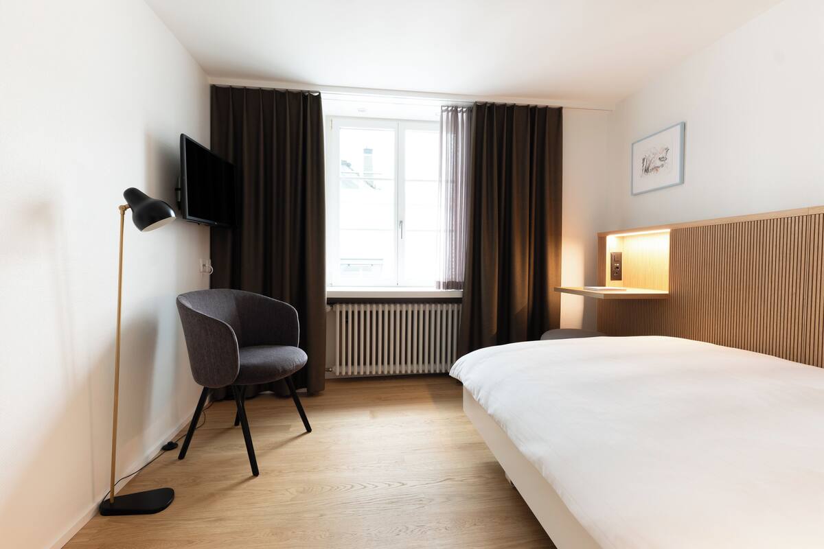 A hotel room with a bed, a chair, a TV, a window with curtains, and a radiator.