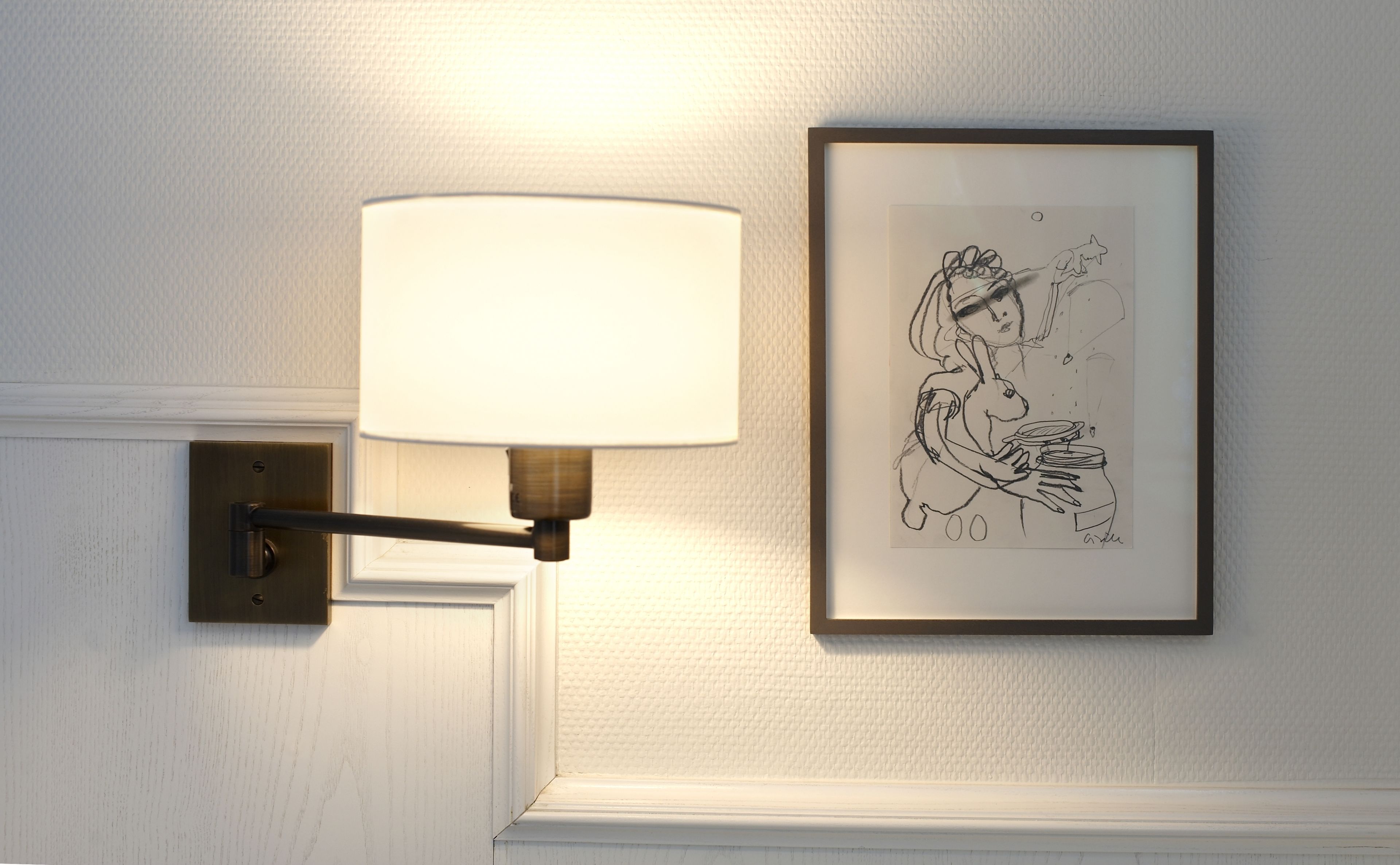 Una lámpara de pared con pantalla blanca y un dibujo enmarco de una mujer sosteniendo un conejo.