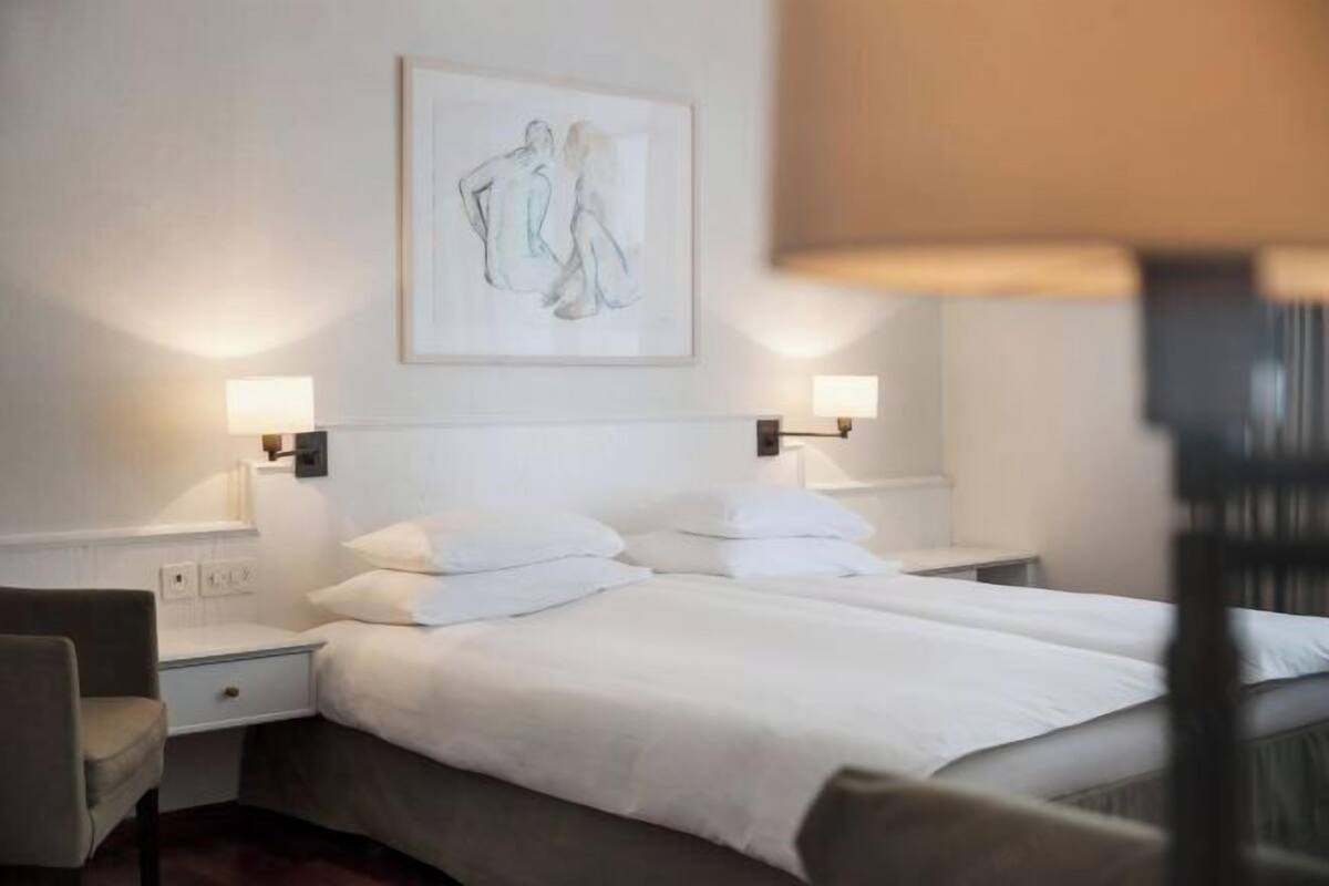 Habitación de hotel con cama doble, dos mesitas de noche, una silla y una obra de arte colgada en la pared.