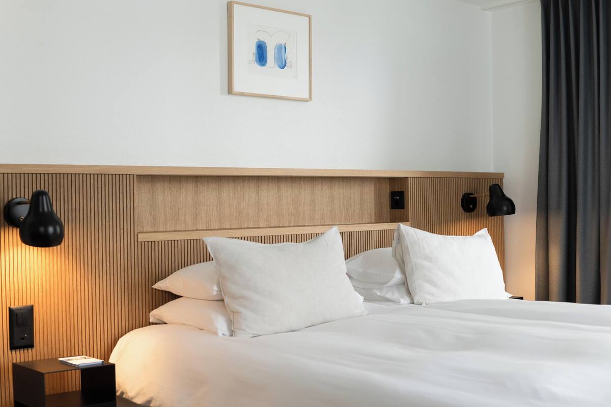 A hotel room with a wooden headboard, white bedding, two black wall sconces, and a framed picture on the wall.