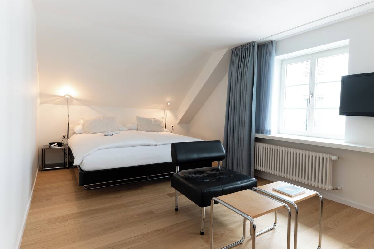 Una habitación de hotel moderna con cama, mesitas de noche, un escritorio, una silla, un televisor y una ventana con cortinas.