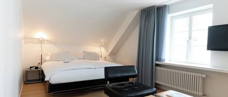 Ein modernes Hotelzimmer mit Bett, Nachttischen, Schreibtisch, Stuhl, Fernseher und einem Fenster mit Vorhängen.