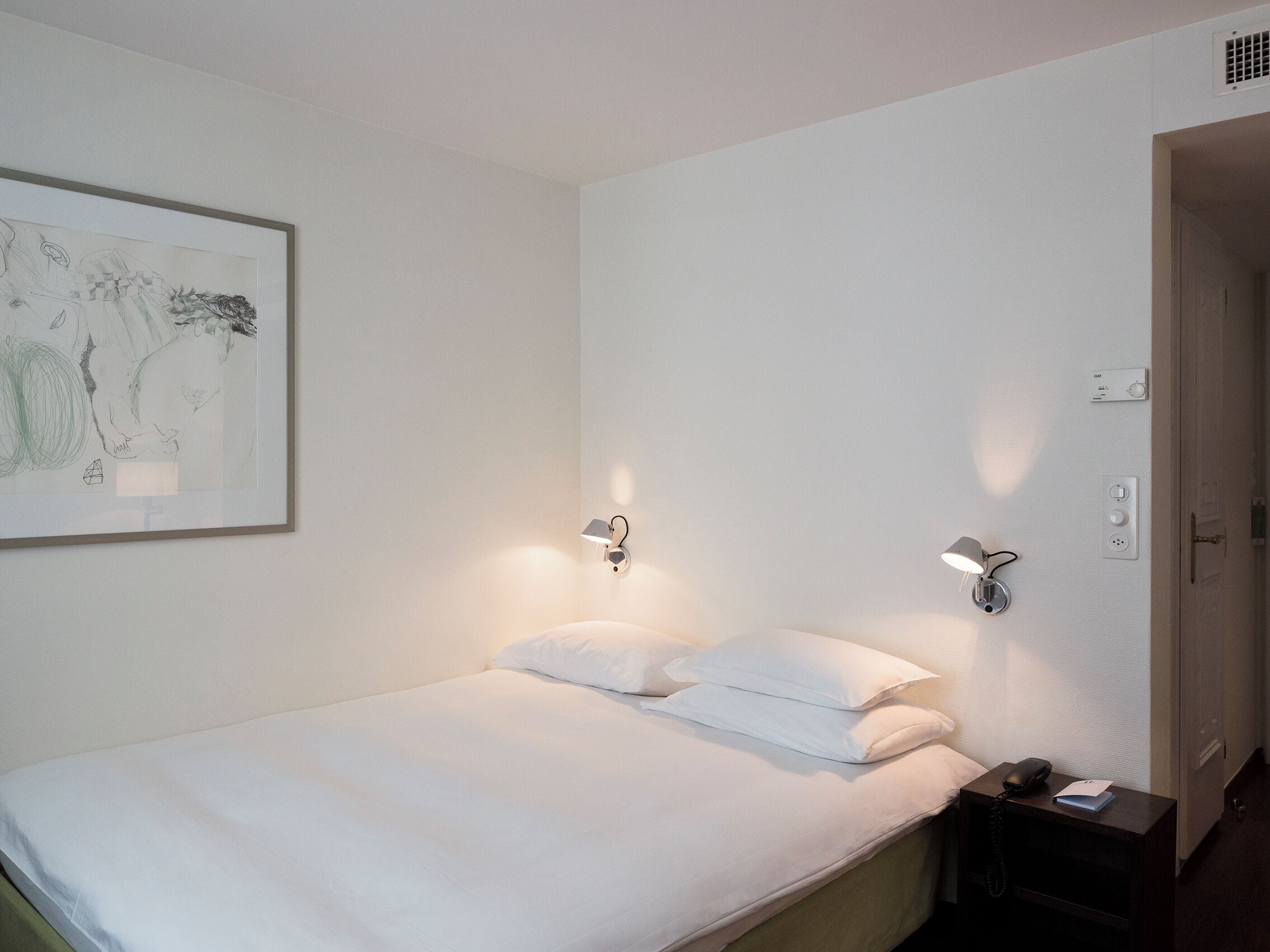 A neatly made bed with white linens, a bedside table with a lamp, and a wall-mounted light fixture.