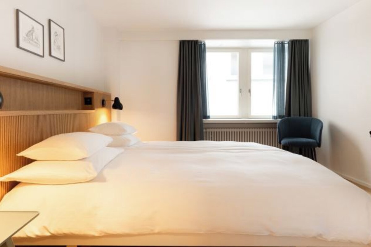 A hotel room with a bed, a chair, a desk, a window with curtains, and framed pictures on the wall.