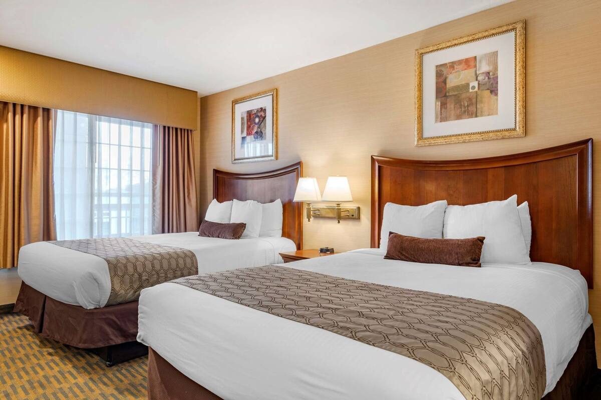 A hotel room with two beds, a large window with curtains, a wall-mounted painting, and a nightstand with lamps.