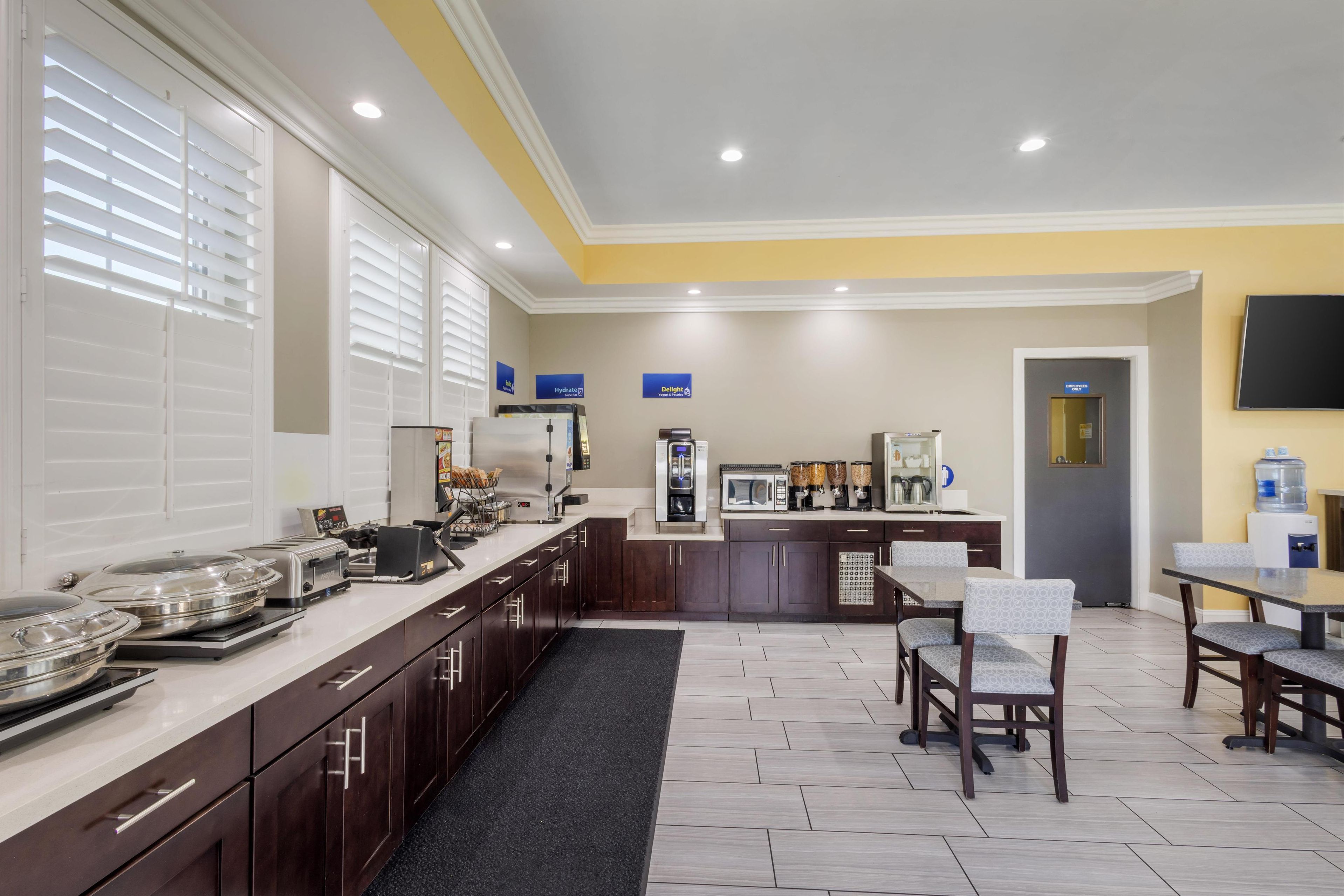 A modern kitchen with a long counter, microwave, coffee machine, and a water dispenser. There are chairs and tables for dining, and a TV mounted on the wall.