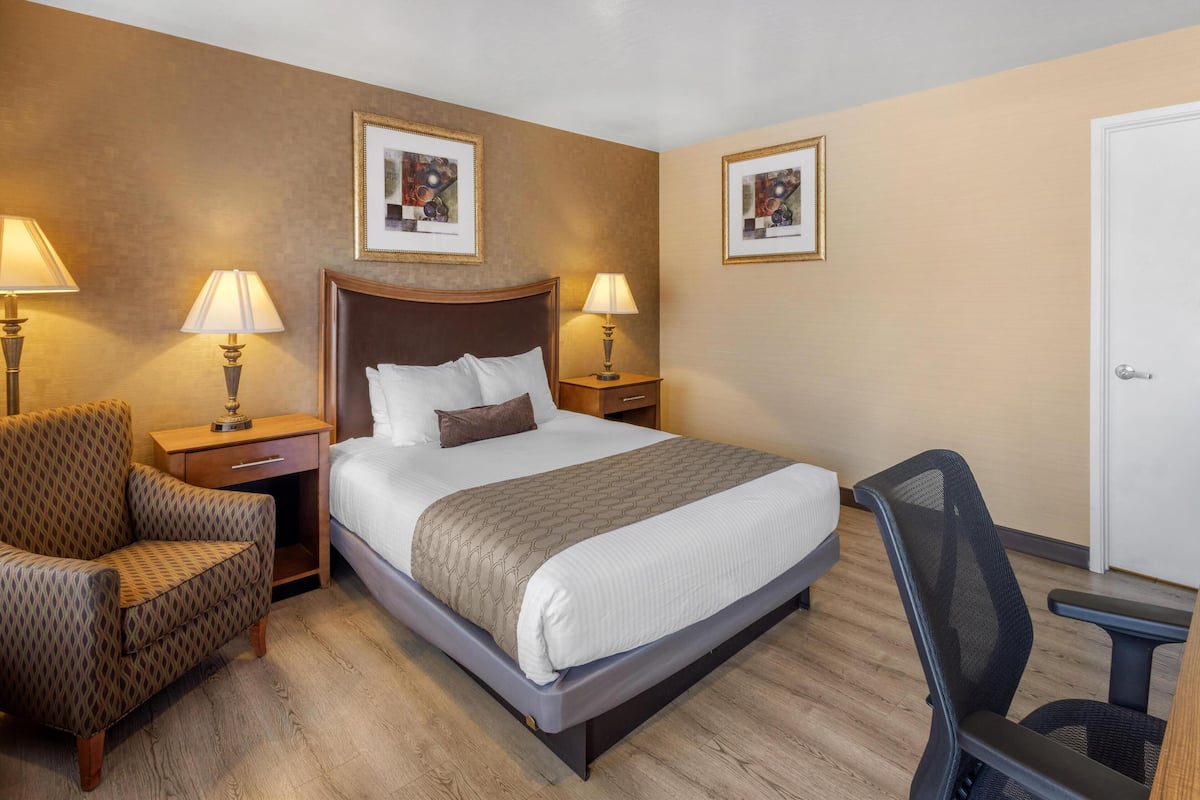 A hotel room with a large bed, two bedside tables with lamps, a chair, a desk, and two framed pictures on the walls.