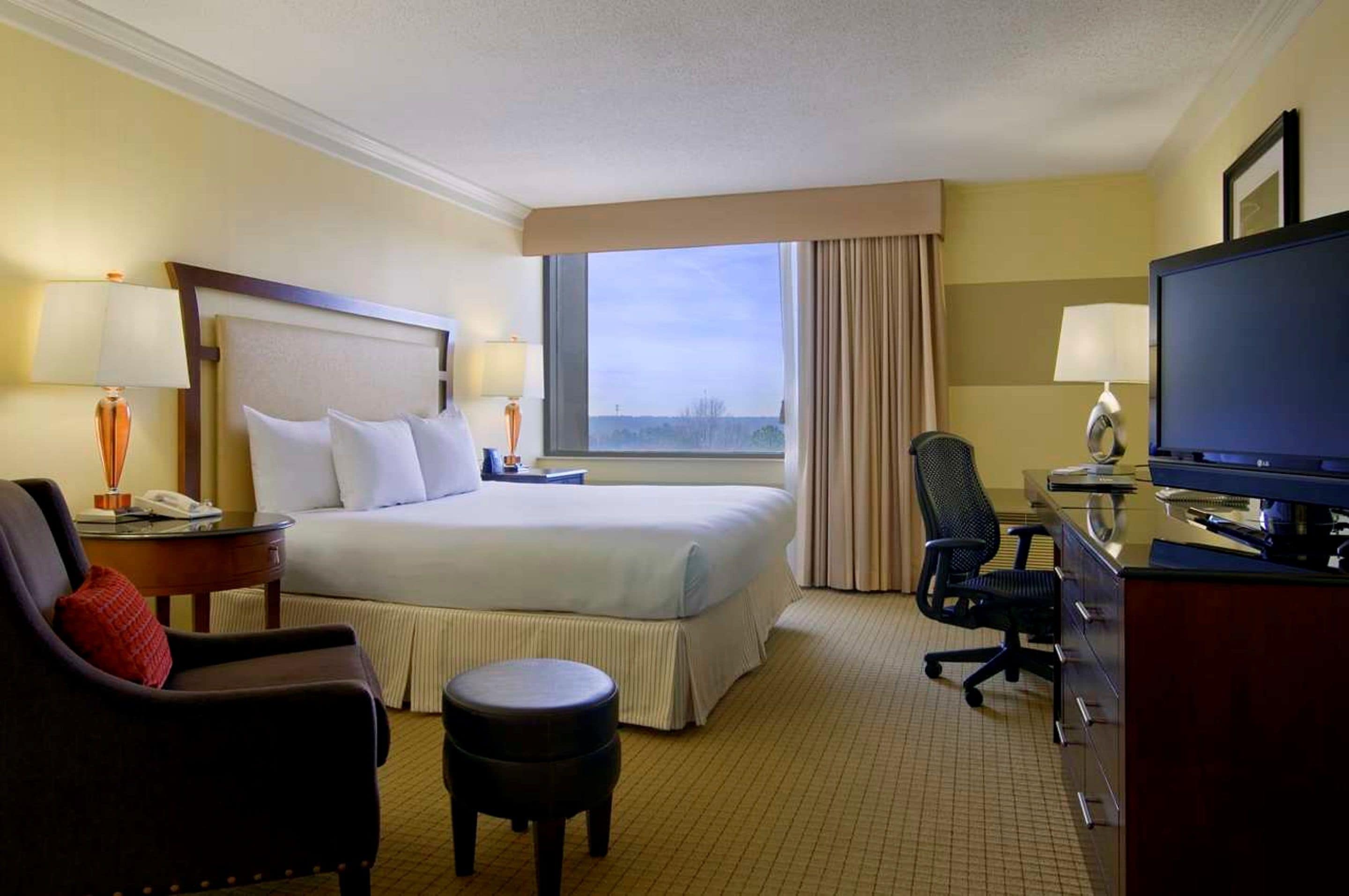 Habitación de hotel con una cama grande, un escritorio con computadora, una silla, un taburete pequeño y un televisor.