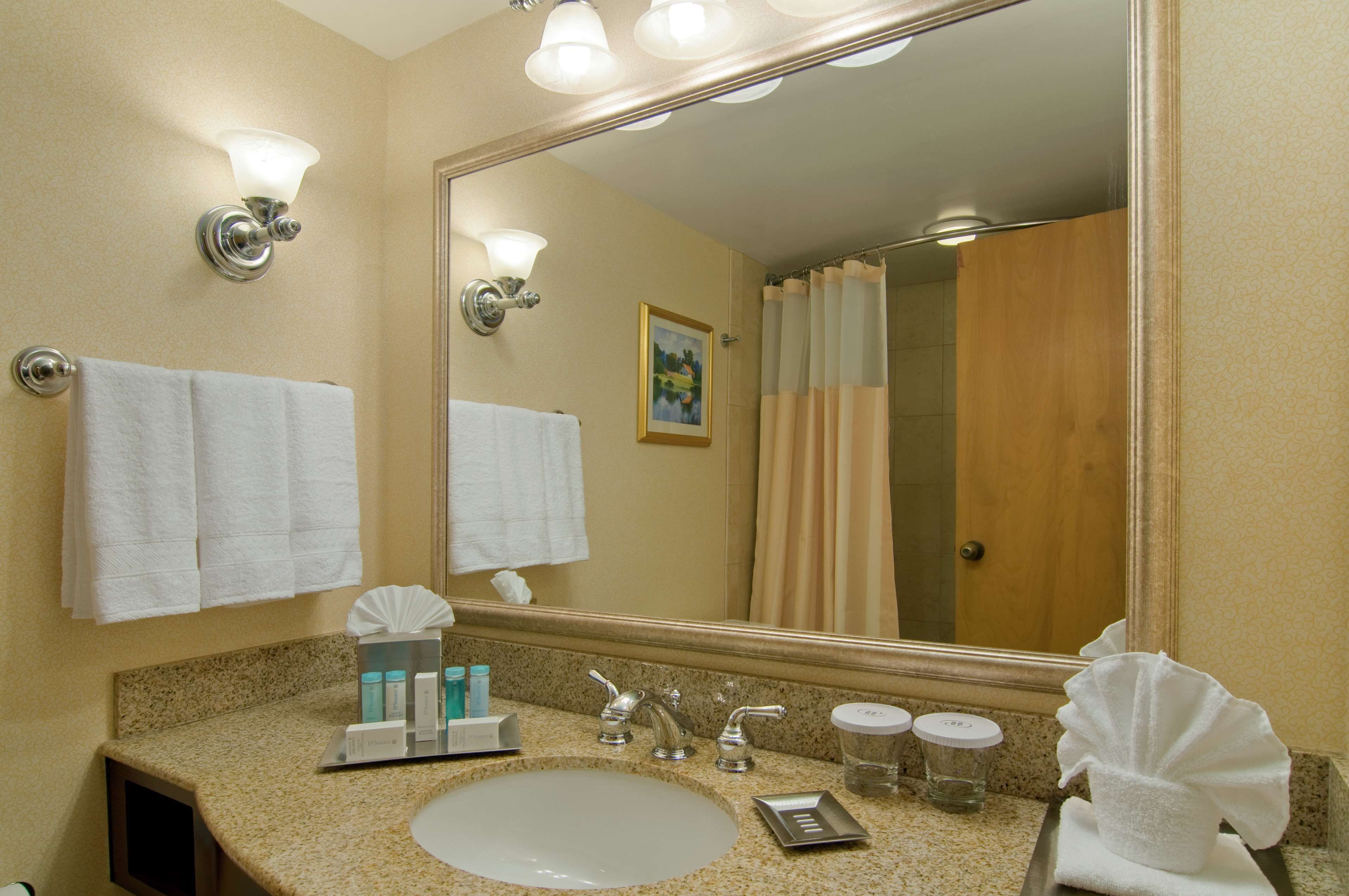 Une salle de bain d’hôtel avec un grand miroir, deux appliques murales, un lavabo avec des articles de toilette et un rideau de douche.