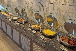 Un bufé con diversos alimentos, entre ellos frutas, cereales y carnes, dispuesto sobre una encimera de mármol con utensilios de metal.