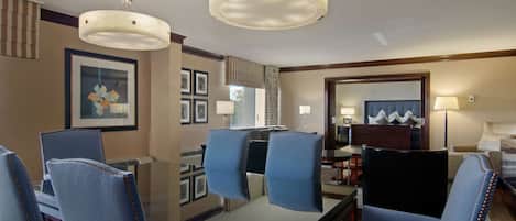 A hotel room with a dining area featuring a glass table and blue upholstered chairs, a large mirror, and a chandelier.