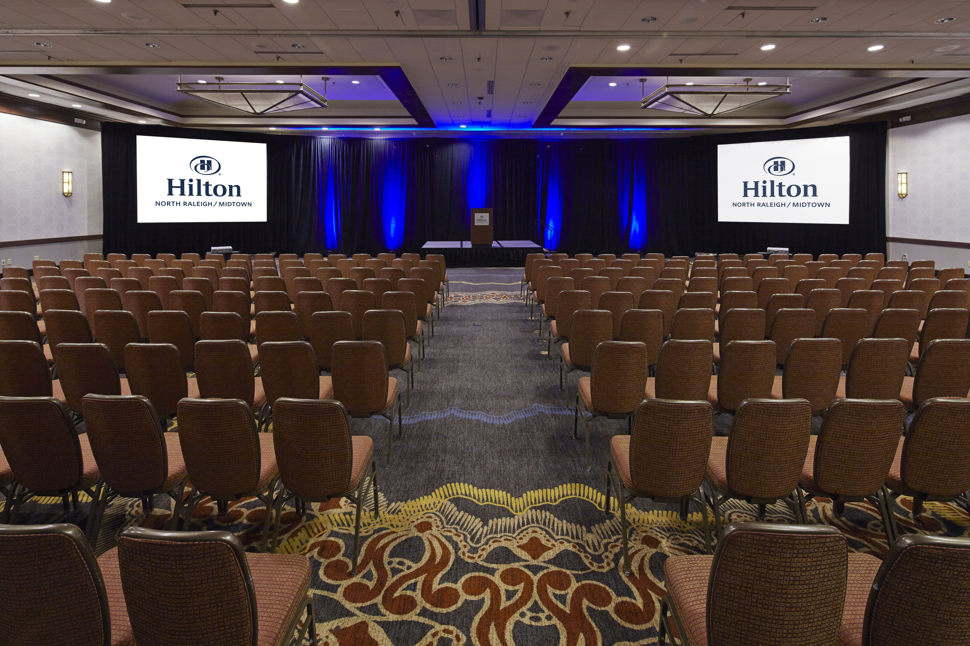 Une salle de conférence avec des rangées de chaises disposées face à une scène sur laquelle figure un grand logo Hilton et l’inscription « North Raleigh/Midtown ».