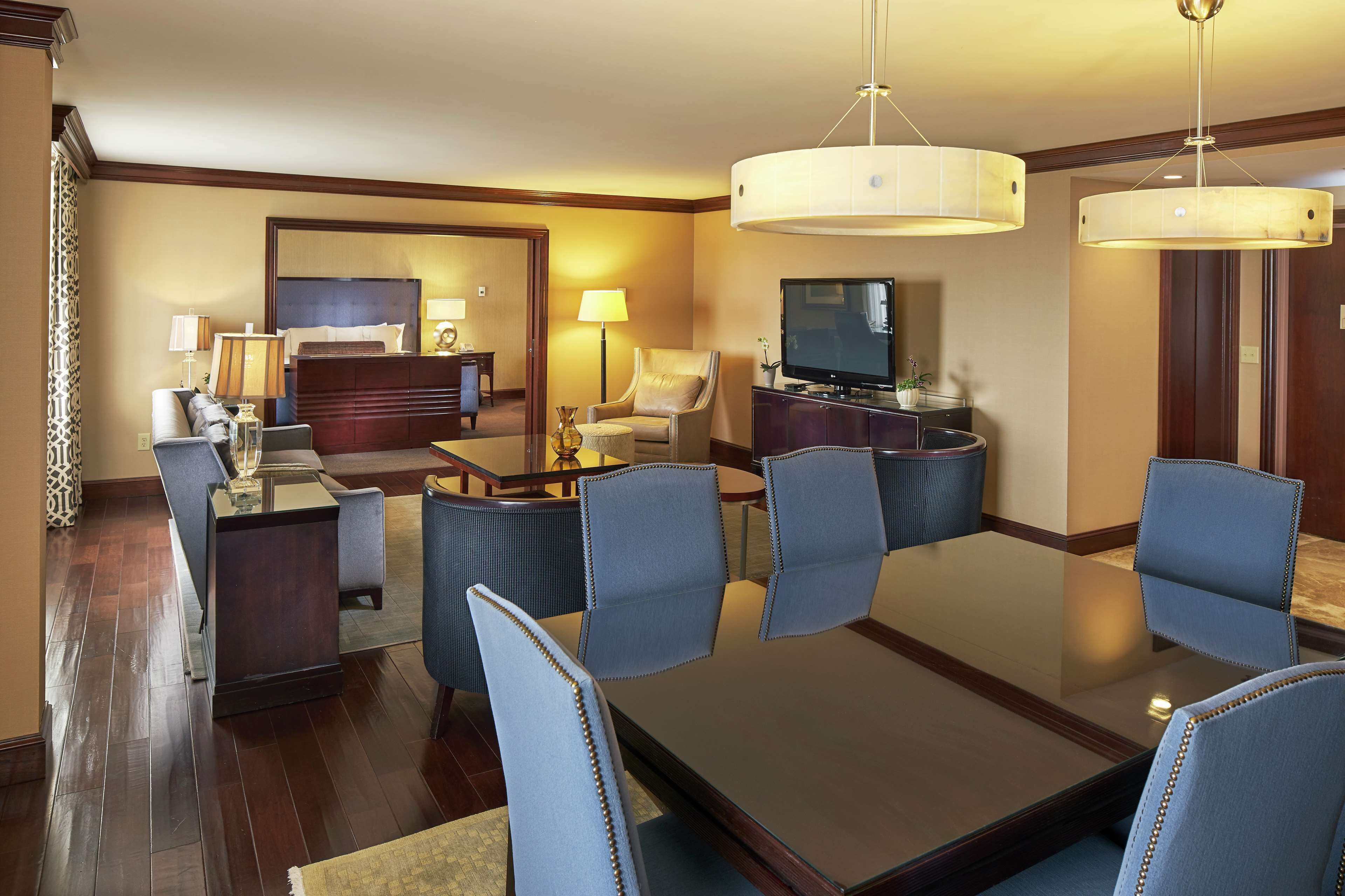 Habitación de hotel con comedor que incluye una mesa grande, sillas, televisor y un rincón de estar con sofá y sillones.