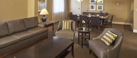 Quarto de hotel com sofá, poltronas, mesa de centro, mesa de jantar e cadeiras, e um candelabro.