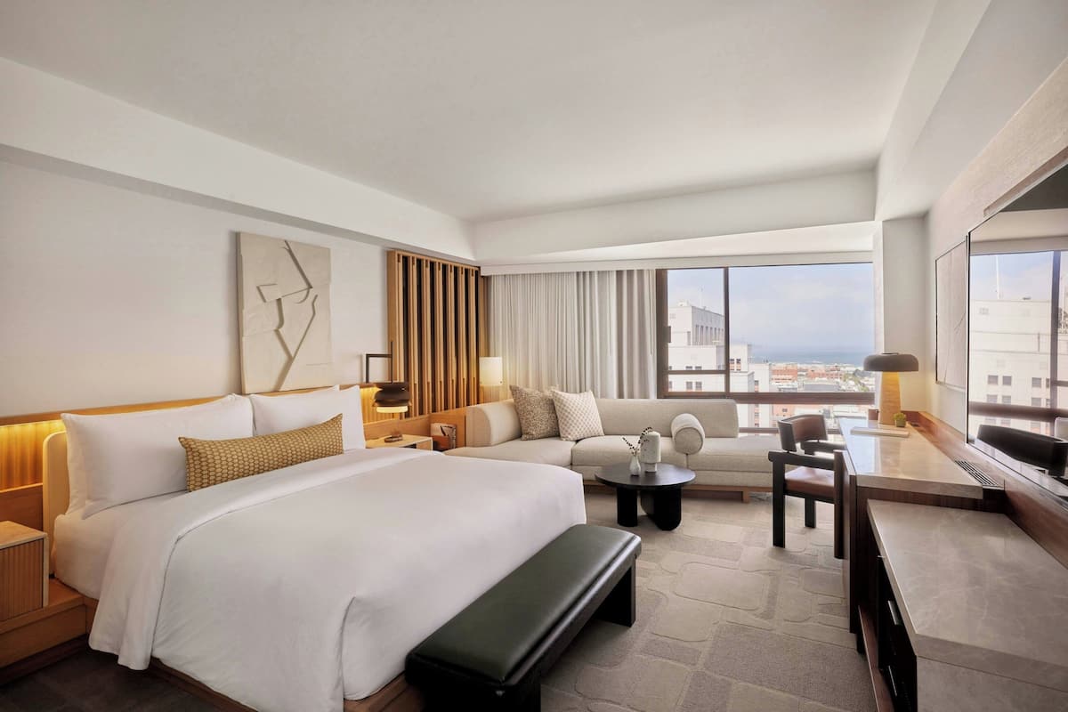 A modern hotel room with a large bed, a sofa, a desk, and a view of the city.