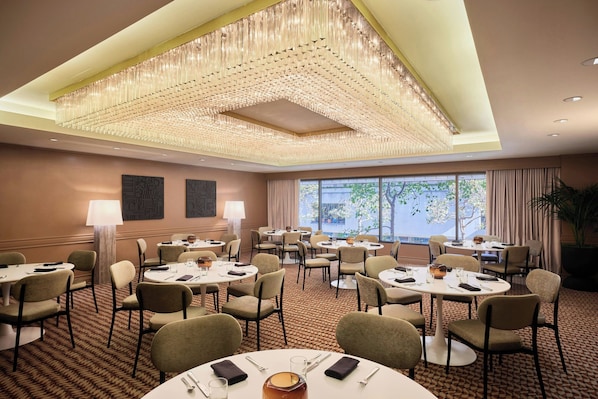 A hotel conference room with round tables, chairs, and a large chandelier.