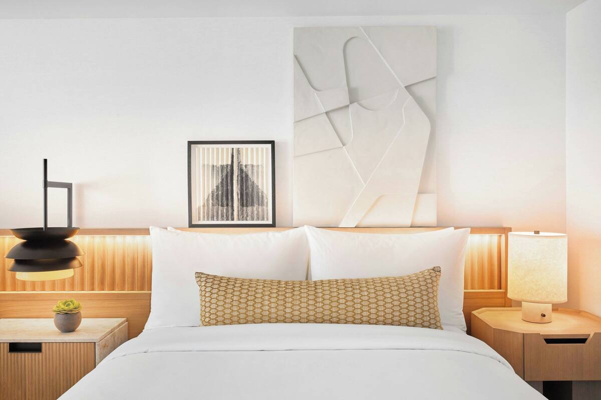 A modern hotel room with a neatly made bed, a wall-mounted abstract art piece, a framed photograph, and a bedside table with a lamp.