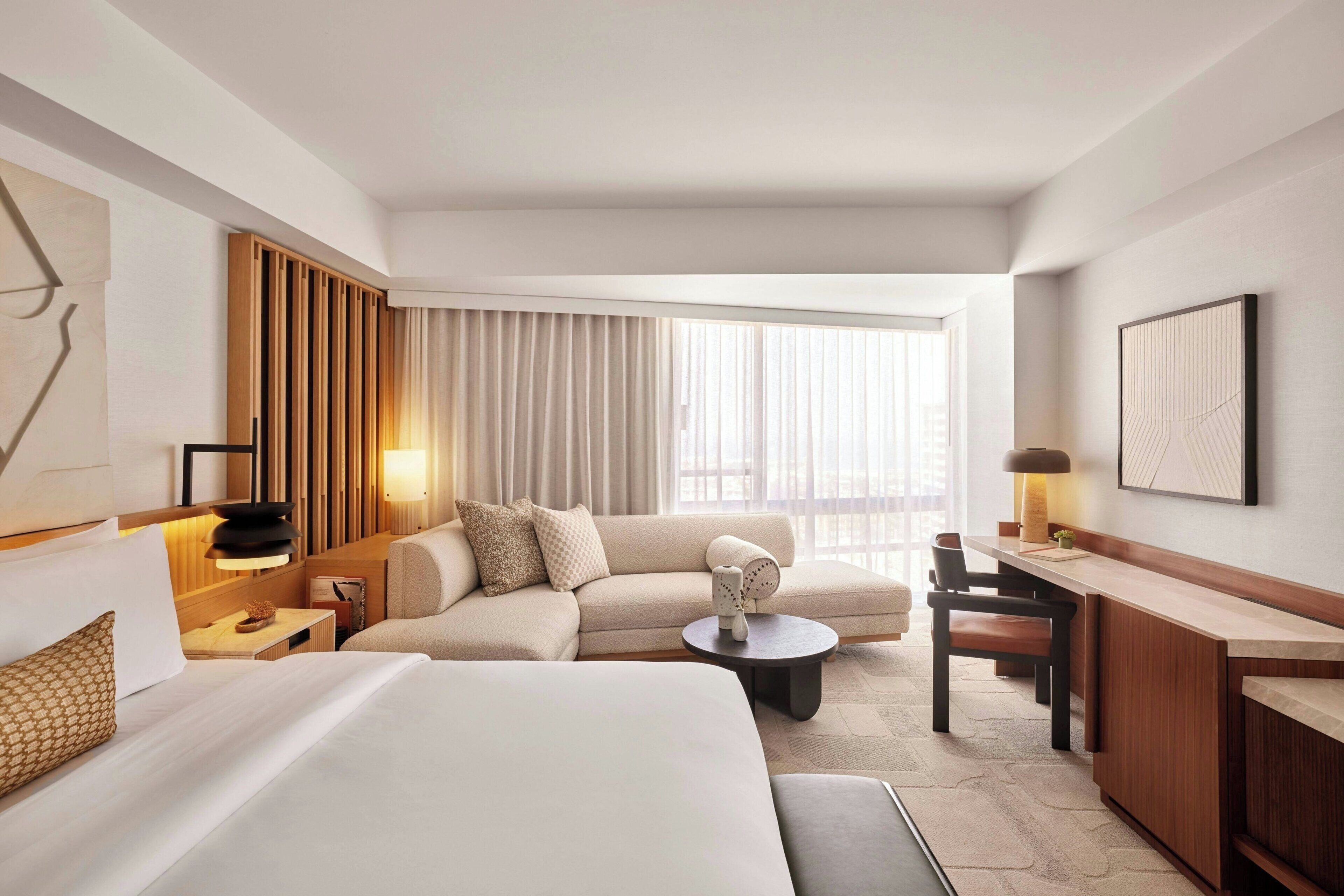 Quarto de hotel moderno com uma cama grande, um sofá, uma escrivaninha e uma mesa de centro. Há uma janela com cortinas, uma luminária e um quadro pendurado na parede.
