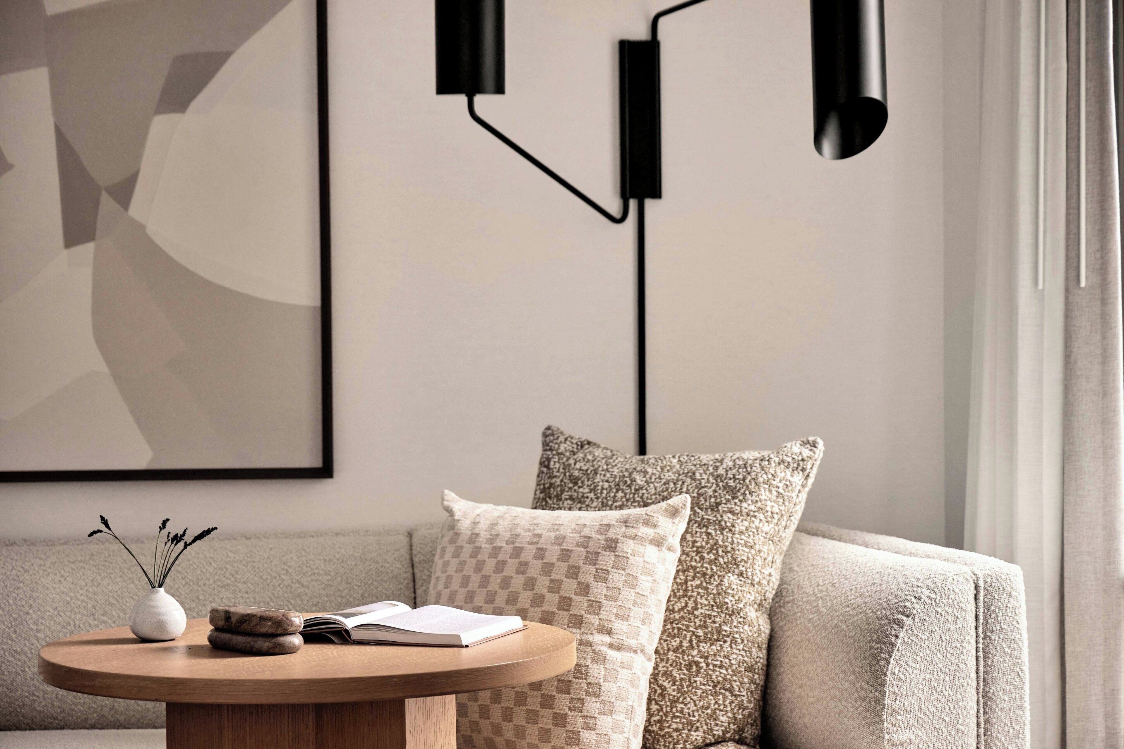 A modern living room with a round wooden coffee table, a vase with dried flowers, and an open book. There are two patterned pillows on a beige sofa, and a framed abstract painting on the wall.