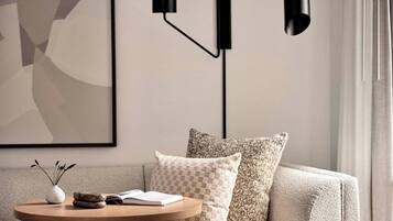 A modern living room with a round wooden coffee table, a vase with dried flowers, and an open book. There are two patterned pillows on a beige sofa, and a framed abstract painting on the wall.