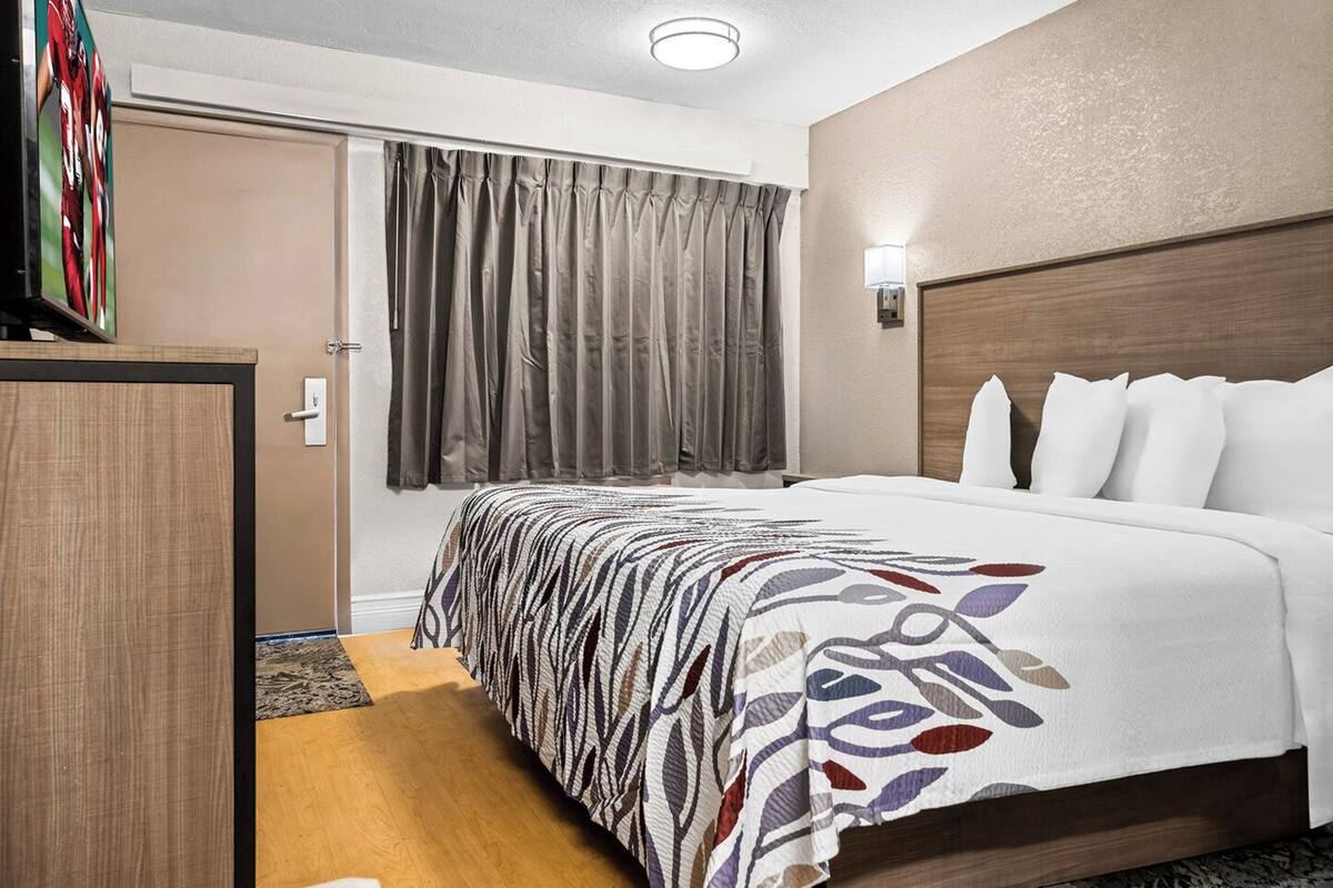 A hotel room with a bed, a wooden headboard, a window with curtains, a bedside table, and a wall-mounted light.