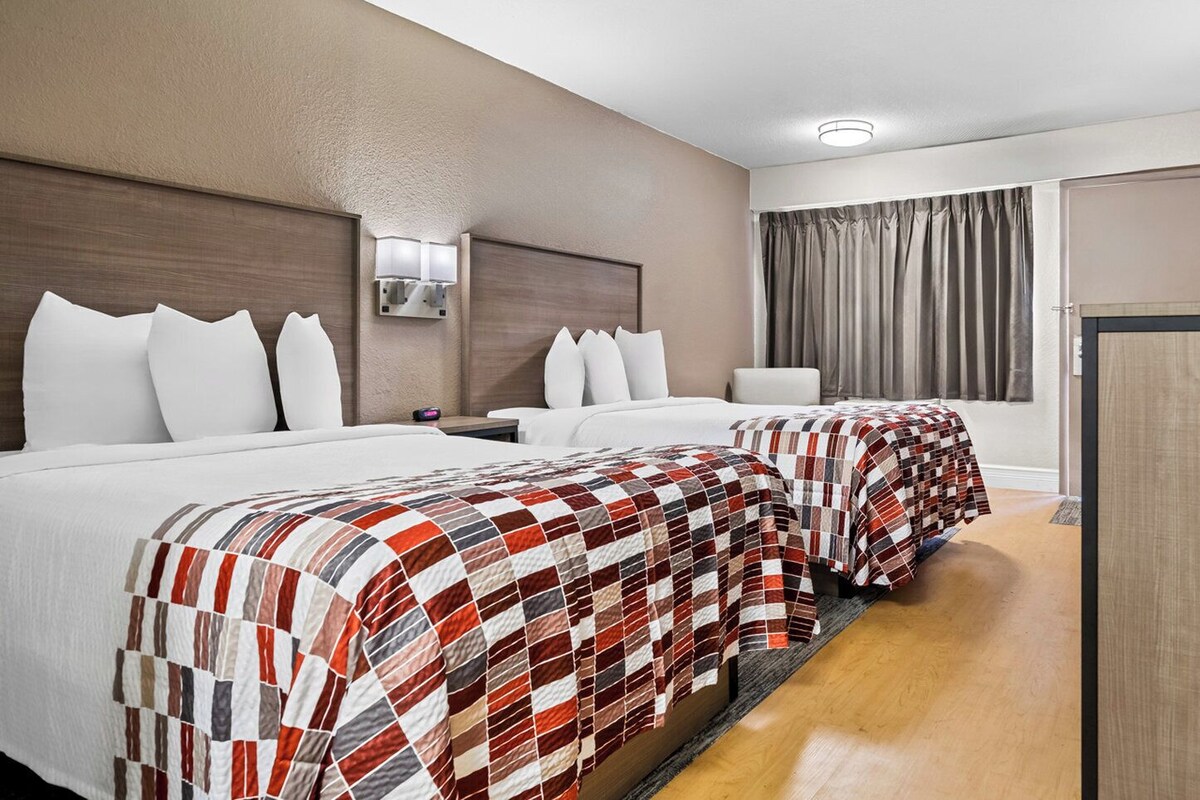 Una habitación de hotel con dos camas, cada una con colcha a cuadros rojo y blanco, almohadas blancas, una mesita de noche con lámpara y una ventana con cortinas.