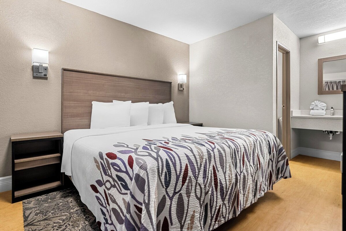 Una habitación de hotel con cama, mesitas de noche, baño con lavamanos y espejo, y una colcha estampada.