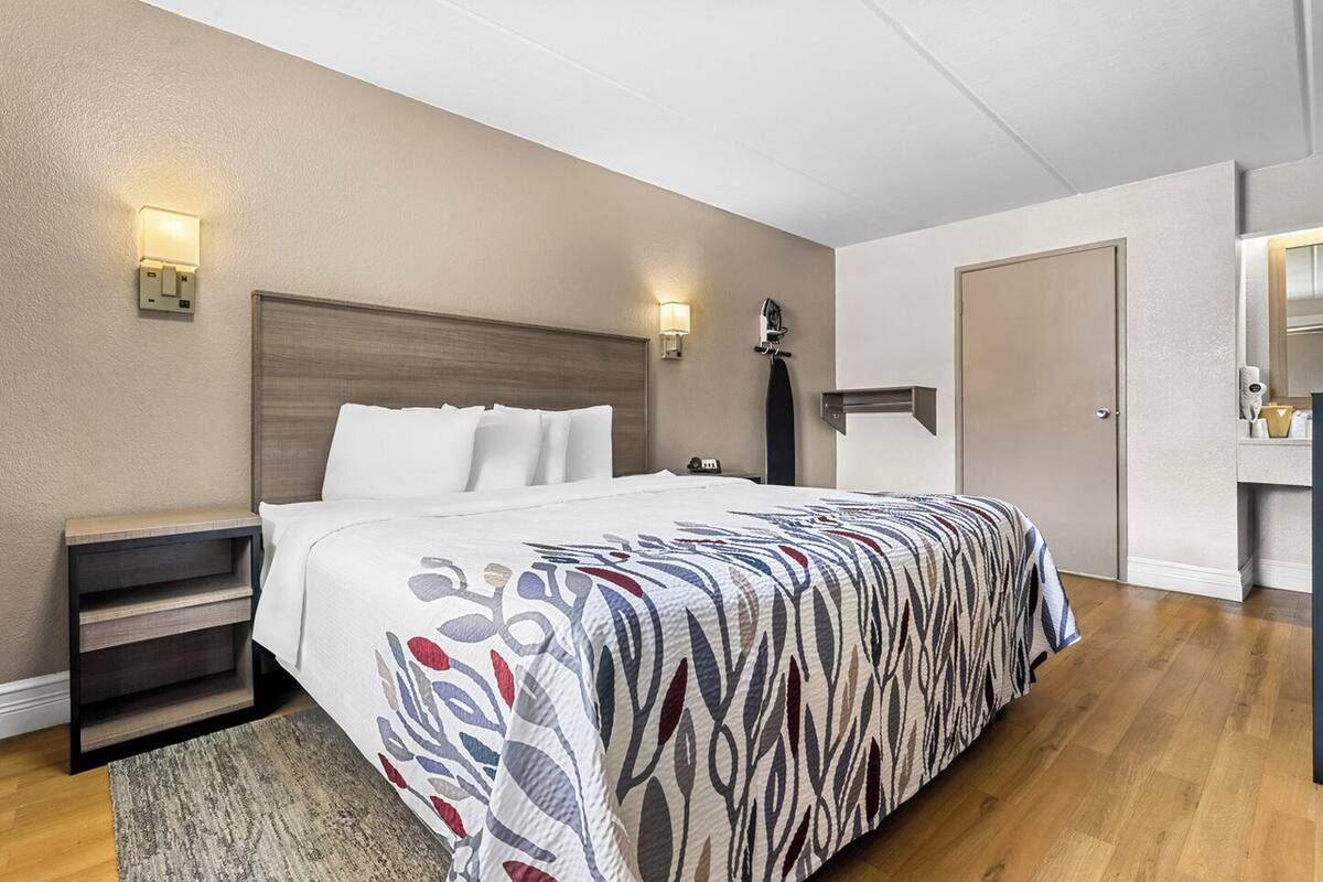 A hotel room with a bed, bedside tables, a nightstand, a wall-mounted lamp, a door, and a small mirror.