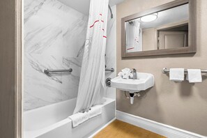 Un baño con pared de ducha de mármol, una bañera blanca, un lavabo con espejo y dos toallas colgadas en un perchero.