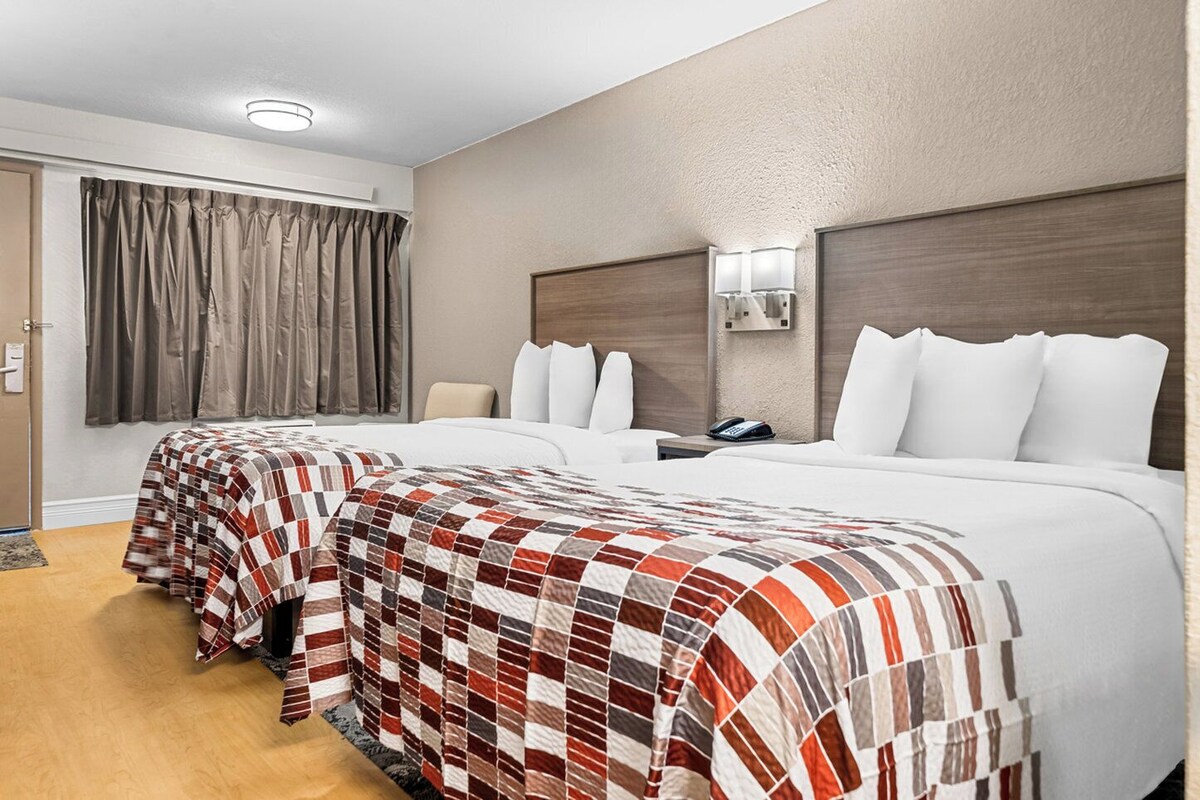 Habitación de hotel con dos camas, cabecero de madera, mesita de noche con lámpara y ventana con cortinas.
