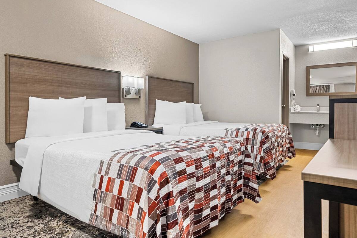 A hotel room with two beds, a nightstand, a desk, a mirror, and a bathroom with a sink and toilet.