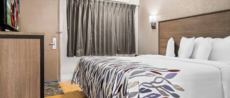 A hotel room with a bed, a wooden headboard, a window with curtains, a bedside table, and a wall-mounted light.