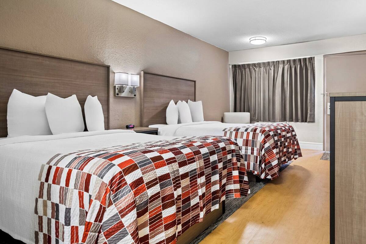 A hotel room with two beds, each with a red and white checkered bedspread, white pillows, a nightstand with a lamp, and a window with curtains.