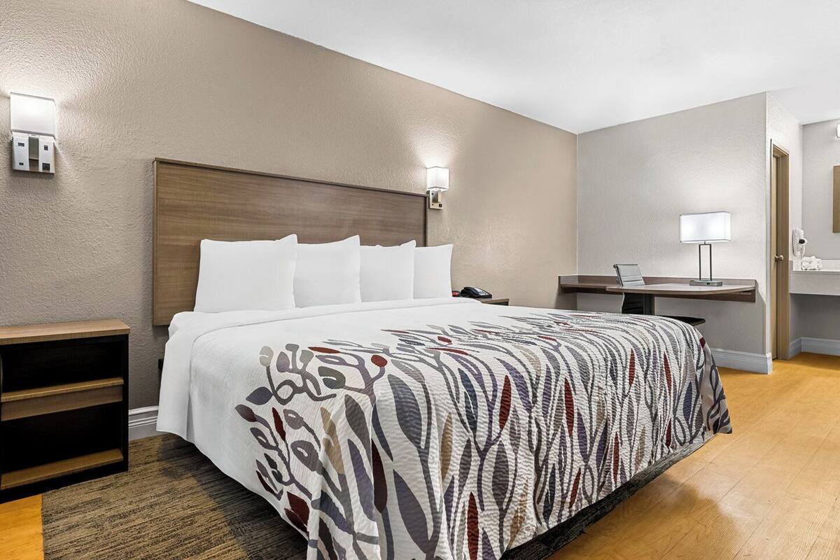 A hotel room with a large bed, a bedside table, a desk with a lamp, and a bathroom visible through an open door.