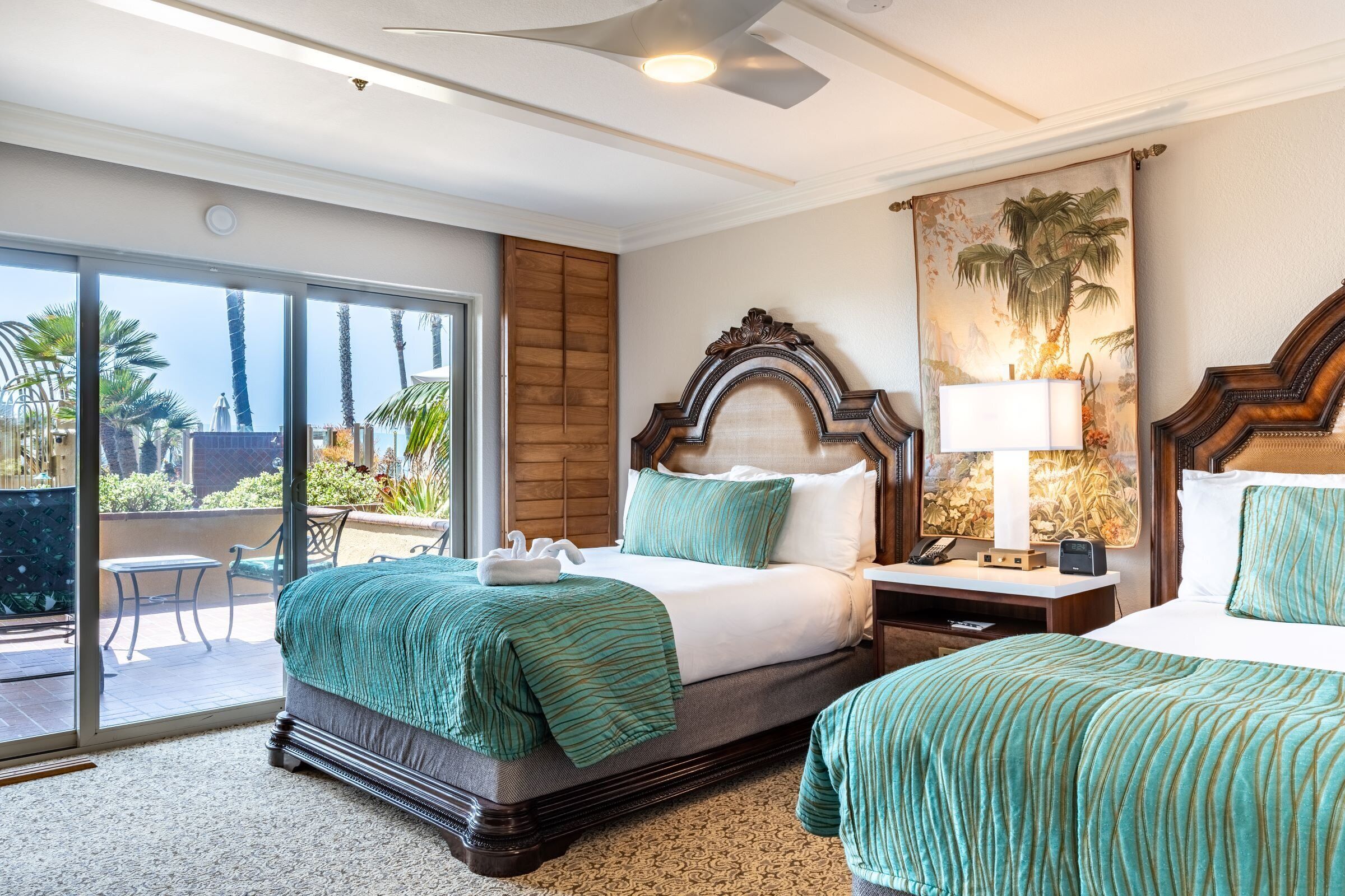 Quarto de hotel com duas camas, uma janela grande com vista para palmeiras e uma varanda.