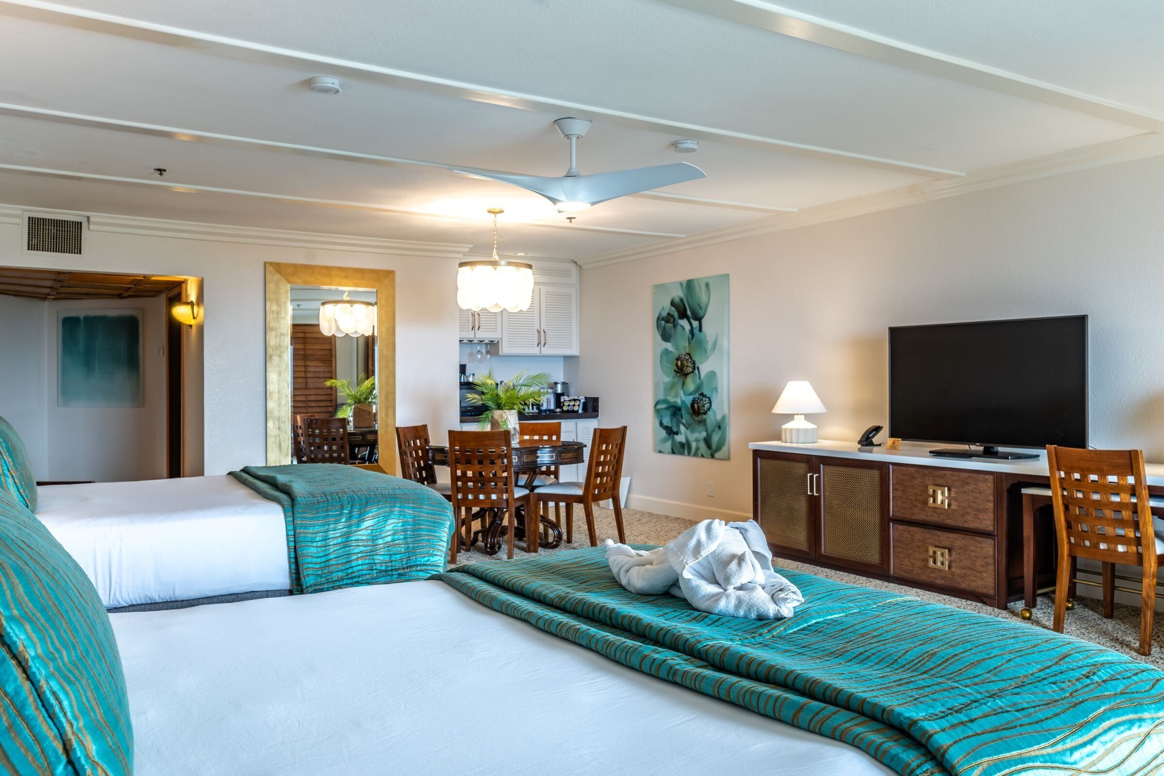 A hotel room with two beds, a desk with a television, a dining area, and a kitchen in the background.