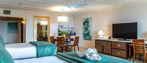 A hotel room with two beds, a desk with a television, a dining area, and a kitchen in the background.