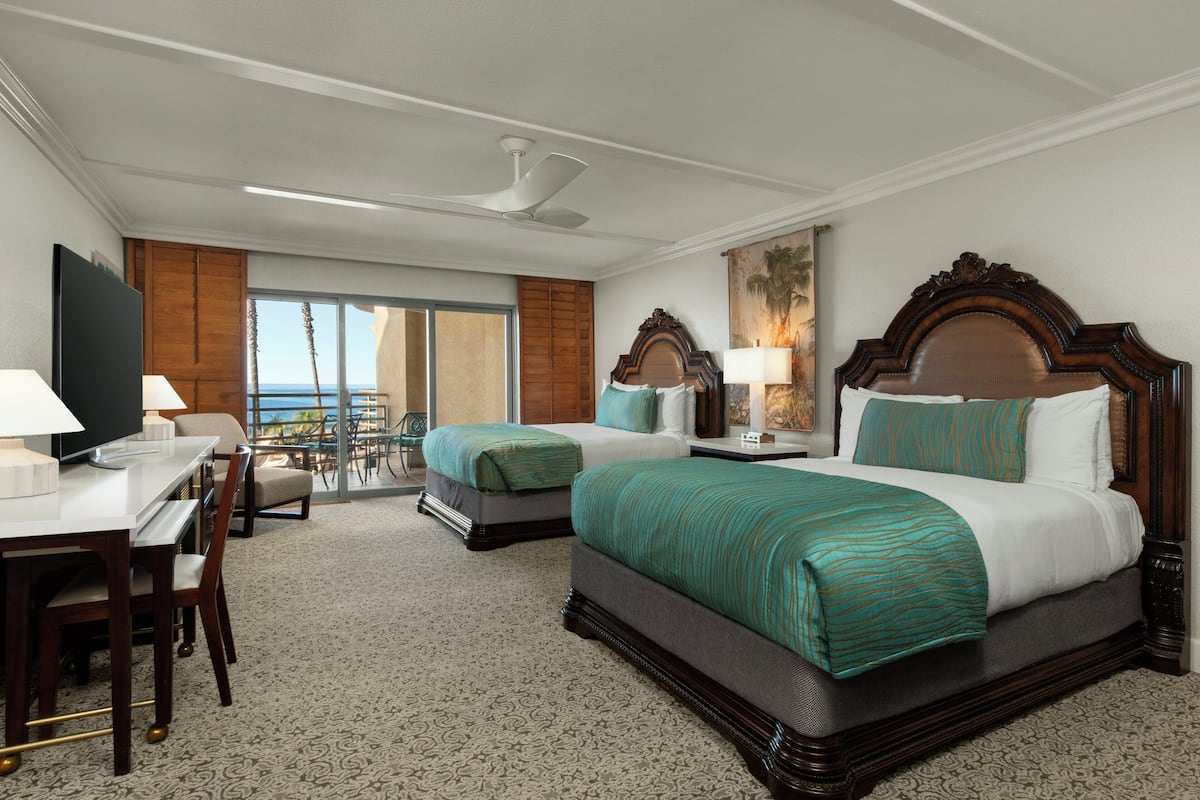 A hotel room with two beds, a desk, and a chair. There is a large window with a view of the ocean.
