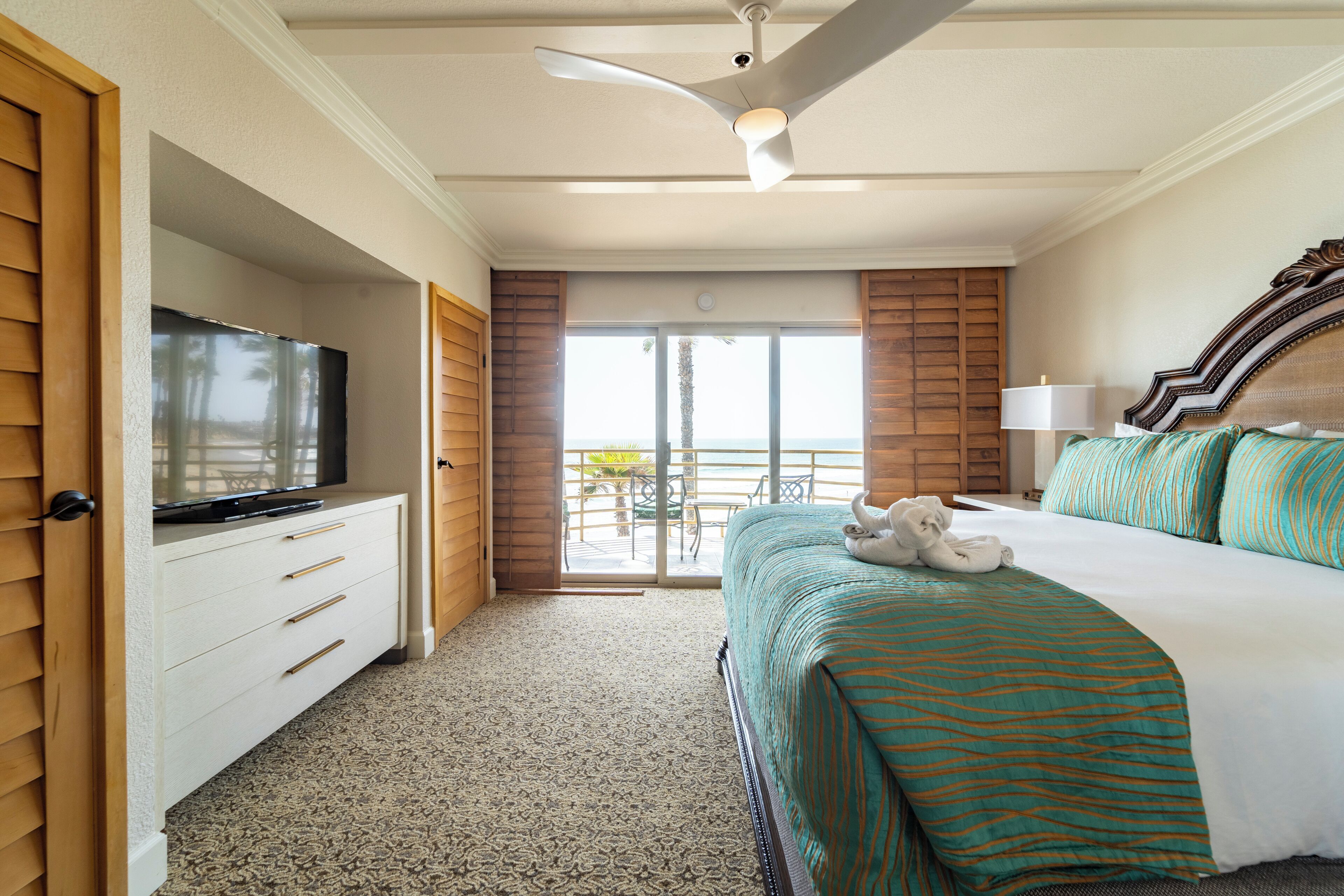 Une chambre d’hôtel avec un lit, un téléviseur posé sur une commode, un ventilateur de plafond et une vue sur l’extérieur.