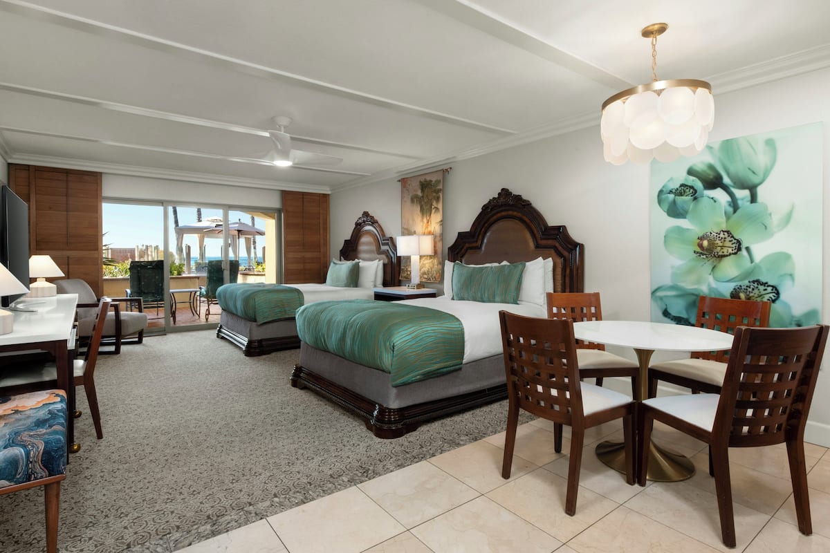 A hotel room with two beds, a dining table, and chairs. There is a view of the outdoors through large windows.